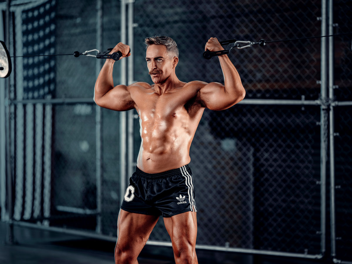 Coach is exercising his arms in a gym, shirtless.